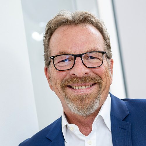 Ein Mann im mittleren Alter mit Brille steht angelehnt an einer Wand. Er trägt ein weißes Hemd und blaues Sakko.