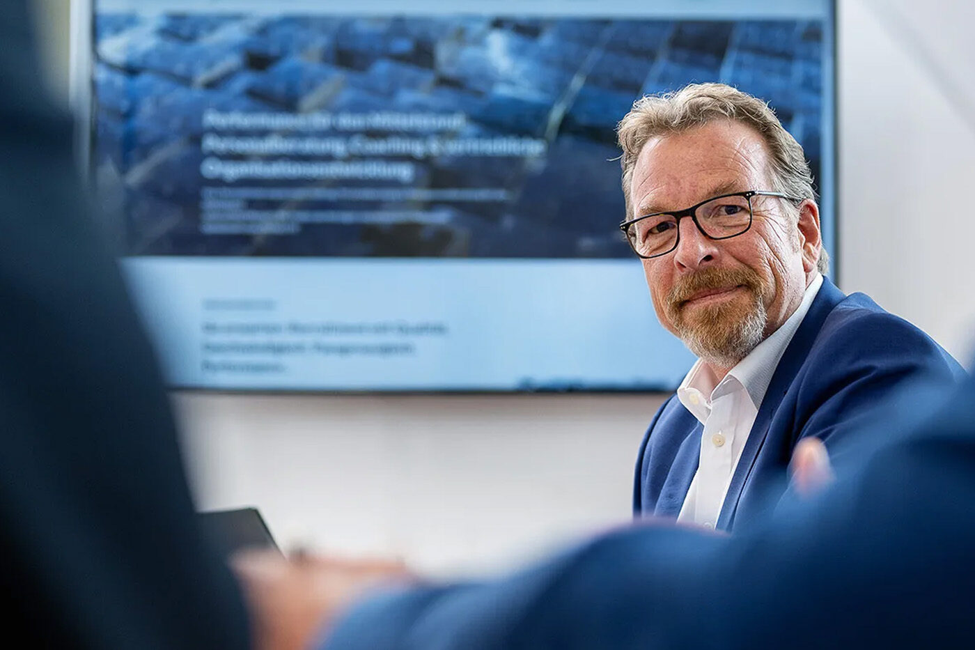 Ein Mann mittleren Alters mit Brille und grauen Haaren schaut rüber und trägt ein blaues Sakko und weißes Hemd. Im Hintergrund ist ein Bildschirm zu sehen.