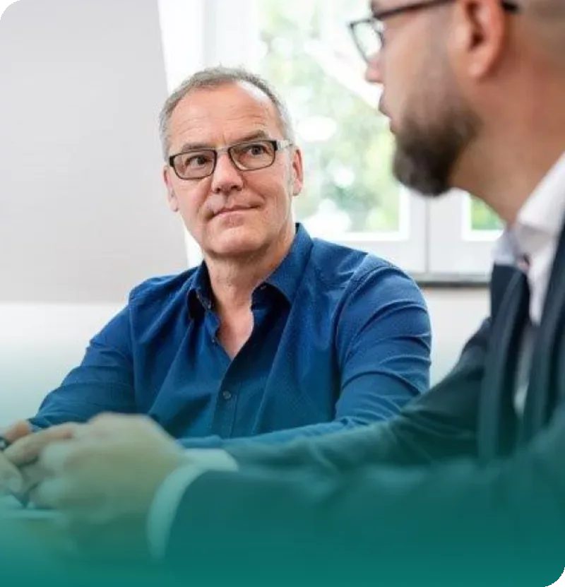 Zwei Männer sitzen an einem Tisch. Der eine hört dem anderen aufmerksam zu. Beide tragen eine Brille und Hemd.